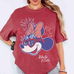 Op de afbeelding: Rood T-shirt met een Minnie Mouse-afbeelding met een hartvormige zonnebril en een ruitjesstrik. Het ontwerp bevat vuurwerk en de tekst "White & Cute". Het shirt heeft korte mouwen en een relaxte pasvorm.