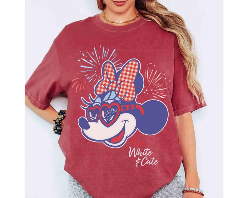 Op de afbeelding: Rood T-shirt met een Minnie Mouse-afbeelding met een hartvormige zonnebril en een ruitjesstrik. Het ontwerp bevat vuurwerk en de tekst "White & Cute". Het shirt heeft korte mouwen en een relaxte pasvorm.