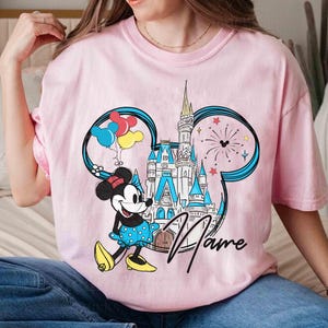 May include: Pink t-shirt with a graphic of Mickey Mouse's head, featuring a castle, balloons, and fireworks. The text "Name" is printed on the shirt.