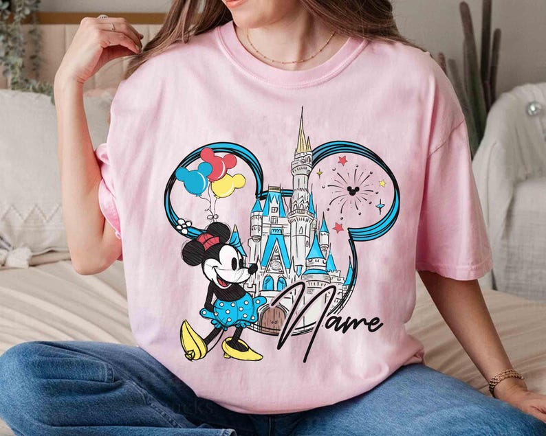 May include: Pink t-shirt with a graphic of Mickey Mouse's head, featuring a castle, balloons, and fireworks. The text "Name" is printed on the shirt.