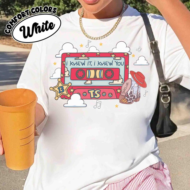 May include: A white t-shirt featuring a graphic print of a red cassette tape with the text 'I KNEW IT, I KNEW YOU' and 'TS'. The design includes a yellow sheriff star with the number 13, a cowboy boot, clouds, stars, and musical notes.
