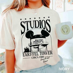 May include: Ivory-colored t-shirt featuring a black graphic of a water tower with Mickey Mouse ears, the text "STUDIOS" above, and "EARFFEL TOWER CIRCA 1989 backlot tour" below. The shirt is paired with a white mug with a yellow smiley face.