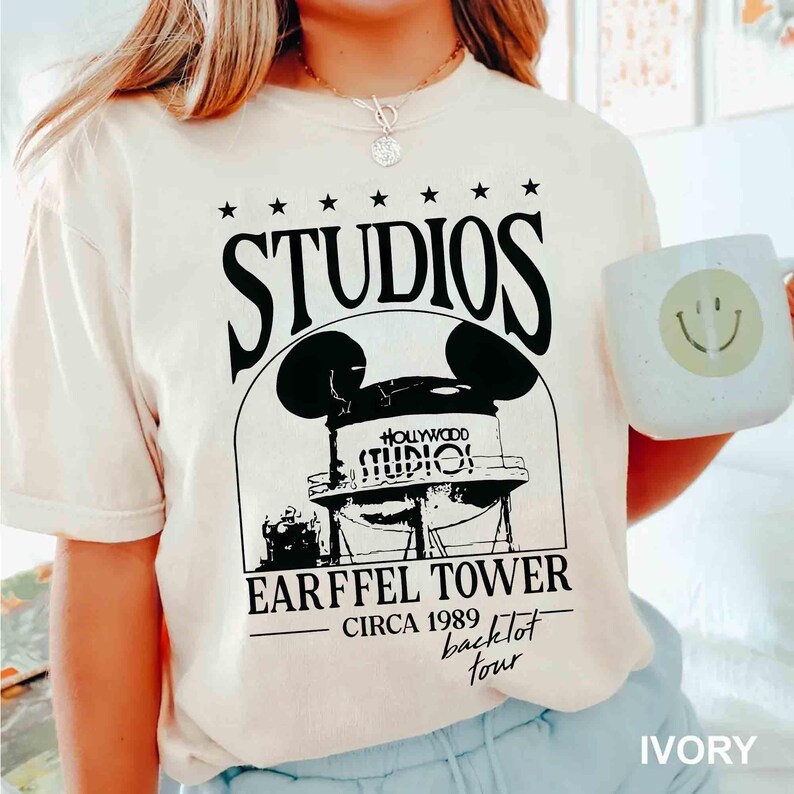 May include: Ivory-colored t-shirt featuring a black graphic of a water tower with Mickey Mouse ears, the text "STUDIOS" above, and "EARFFEL TOWER CIRCA 1989 backlot tour" below. The shirt is paired with a white mug with a yellow smiley face.