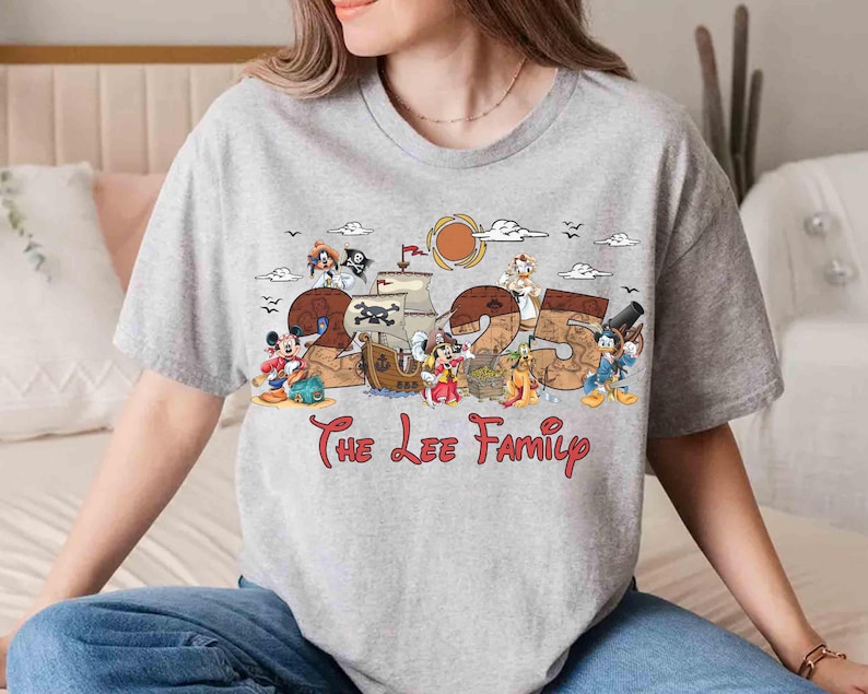 May include: A grey t-shirt with a pirate-themed design, the number "25", and cartoon characters. The text "The Lee Family" is written in red script below the design. The shirt has short sleeves.