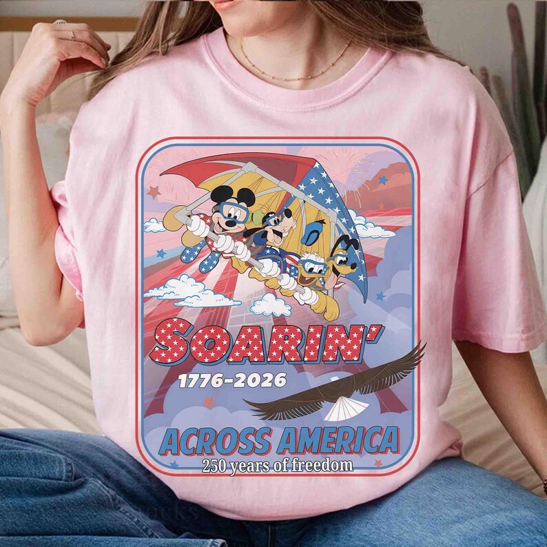 May include: Pink t-shirt featuring a graphic of Mickey Mouse, Goofy, Donald Duck, and other characters soaring through the sky. The design includes the text "Soarin' 1776-2026 Across America 250 years of freedom" and an eagle.