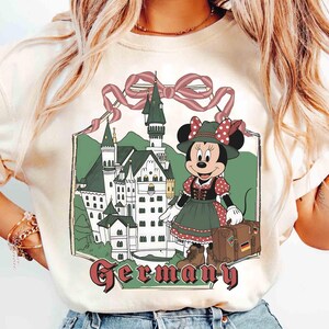 May include: Cream-colored t-shirt featuring a graphic of Minnie Mouse in traditional German attire, standing in front of a castle. The word "Germany" is written in a burgundy font below the image. A pink ribbon tops the castle.