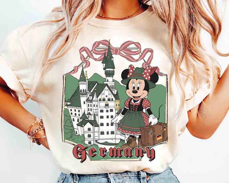 May include: Cream-colored t-shirt featuring a graphic of Minnie Mouse in traditional German attire, standing in front of a castle. The word "Germany" is written in a burgundy font below the image. A pink ribbon tops the castle.