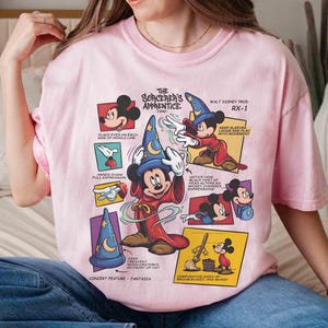 May include: A light pink t-shirt featuring a graphic of Mickey Mouse as the Sorcerer's Apprentice. The design includes multiple panels with illustrations and text, including "The Sorcerer's Apprentice 1940" and "Walt Disney Prod RX-1".