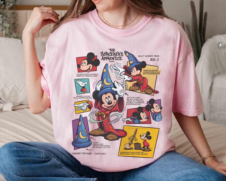May include: A light pink t-shirt featuring a graphic of Mickey Mouse as the Sorcerer's Apprentice. The design includes multiple panels with illustrations and text, including "The Sorcerer's Apprentice 1940" and "Walt Disney Prod RX-1".