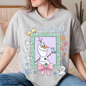 May include: A heather gray t-shirt featuring a graphic of a snowman character inside a decorative green frame. The design includes a gold crown, a small blue lizard, a pink bow, falling autumn leaves, blue snowflakes, and several small snowmen.