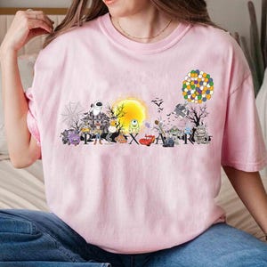 May include: A light pink t-shirt featuring a colorful graphic design with the word "PIXAR" and various cartoon characters. The design includes a yellow sun, a haunted house, and a cluster of balloons.