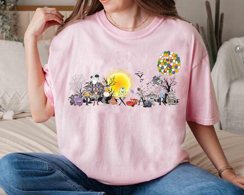 May include: A light pink t-shirt featuring a colorful graphic design with the word "PIXAR" and various cartoon characters. The design includes a yellow sun, a haunted house, and a cluster of balloons.