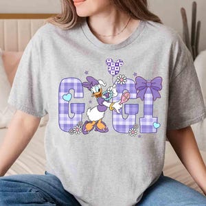 May include: A light gray t-shirt featuring the word "GGL" in a purple plaid pattern. The letter "G" has a blue heart, and the letter "L" has a purple bow. Daisy Duck is holding a balloon and a hairbrush.