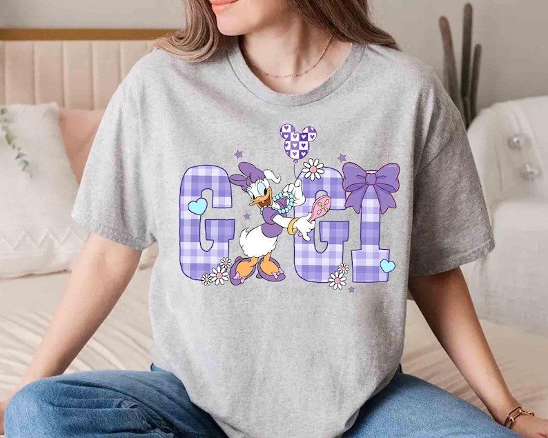 May include: A light gray t-shirt featuring the word "GGL" in a purple plaid pattern. The letter "G" has a blue heart, and the letter "L" has a purple bow. Daisy Duck is holding a balloon and a hairbrush.