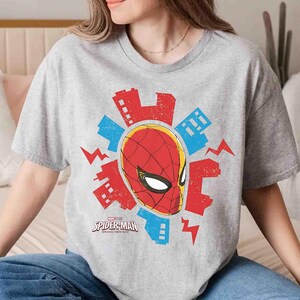 Puede incluir: Una camiseta gris jaspeado de manga corta con un gr&aacute;fico de la m&aacute;scara de Spider-Man rodeada de edificios urbanos estilizados en rojo y azul y rayos. El texto Spider-Man Brand New Day est&aacute; impreso en negro debajo del gr&aacute;fico central.