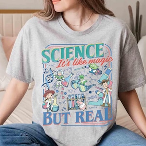 Pu&ograve; includere: Una maglietta grigia con una grafica colorata. Il design presenta il testo "SCIENCE It's like magic BUT REAL" e illustrazioni di personaggi dei cartoni animati e attrezzature scientifiche, come becher e microscopi.