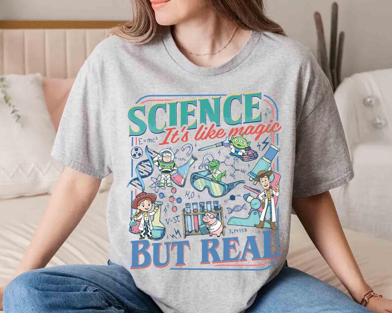 Pu&ograve; includere: Una maglietta grigia con una grafica colorata. Il design presenta il testo "SCIENCE It's like magic BUT REAL" e illustrazioni di personaggi dei cartoni animati e attrezzature scientifiche, come becher e microscopi.