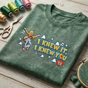 May include: A mineral-washed olive green t-shirt featuring an embroidered graphic of a person playing a guitar, accompanied by the text "I KNEW IT, I KNEW YOU" and "TS" inside a red heart, surrounded by small white stars, clouds, and music notes.