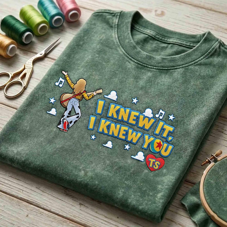 May include: A mineral-washed olive green t-shirt featuring an embroidered graphic of a person playing a guitar, accompanied by the text "I KNEW IT, I KNEW YOU" and "TS" inside a red heart, surrounded by small white stars, clouds, and music notes.