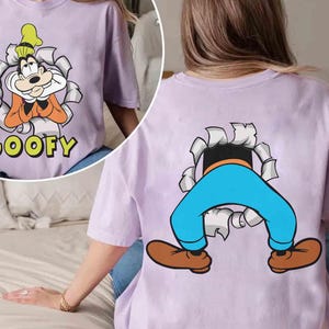 May include: A lilac-colored t-shirt featuring a cartoon image of Goofy. The front of the shirt shows Goofy bursting through a torn paper effect, with the word "GOOFY" below. The back of the shirt shows Goofy's legs and feet.