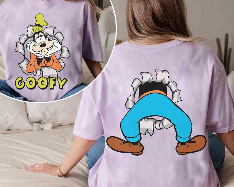 May include: A lilac-colored t-shirt featuring a cartoon image of Goofy. The front of the shirt shows Goofy bursting through a torn paper effect, with the word "GOOFY" below. The back of the shirt shows Goofy's legs and feet.