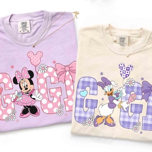 May include: Two Comfort Colors t-shirts, one lavender and one cream, featuring Disney characters. The lavender shirt has Minnie Mouse with the word "GIGI" in pink polka dots. The cream shirt features Daisy Duck with the word "GIGI" in purple plaid.