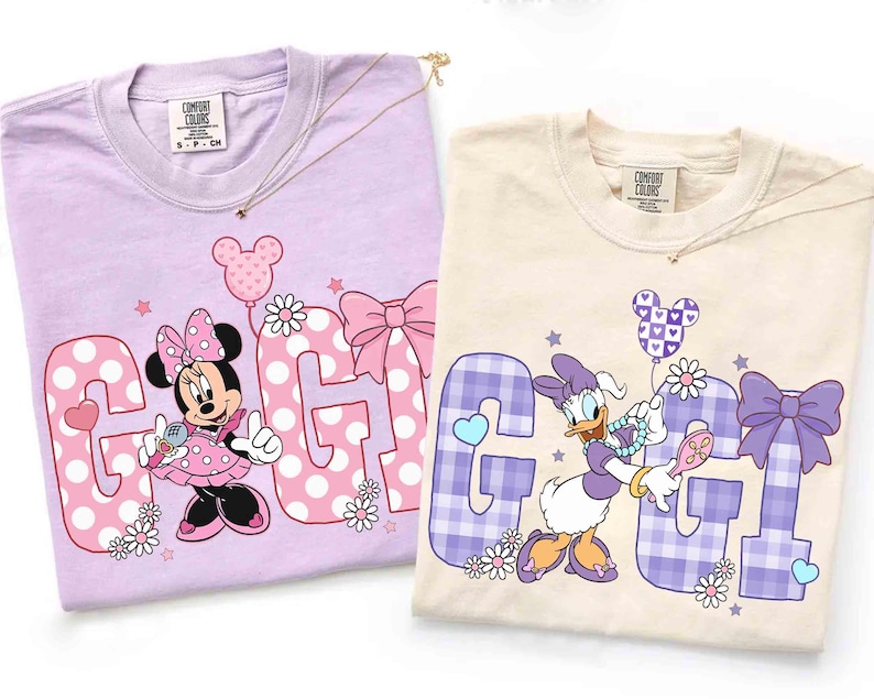 May include: Two Comfort Colors t-shirts, one lavender and one cream, featuring Disney characters. The lavender shirt has Minnie Mouse with the word "GIGI" in pink polka dots. The cream shirt features Daisy Duck with the word "GIGI" in purple plaid.