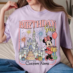 May include: A lilac-colored t-shirt featuring a colorful graphic of Minnie Mouse holding a gift box in front of a castle, fireworks, and a birthday cake. The text "BIRTHDAY Girl" and "Custom Name" are also displayed.