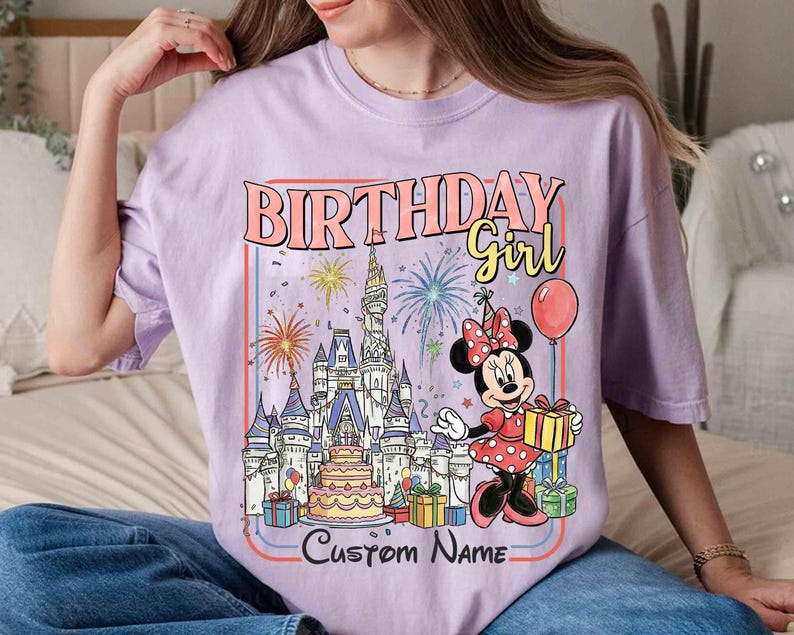 May include: A lilac-colored t-shirt featuring a colorful graphic of Minnie Mouse holding a gift box in front of a castle, fireworks, and a birthday cake. The text "BIRTHDAY Girl" and "Custom Name" are also displayed.