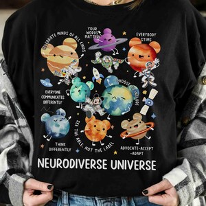 Op de afbeelding: Zwart T-shirt met een kleurrijke afbeelding van planeten met cartoongezichten en de tekst "Celebrate Minds of All Kinds", "Your Words Matter", "Everybody Stims", "Everyone Communicates Differently", "Choose to Include", "See the Able, Not the Label", "Think Differently", "Advocate-Accept-Adapt" en "Neurodiverse Universe".