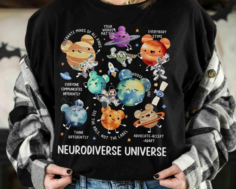 Op de afbeelding: Zwart T-shirt met een kleurrijke afbeelding van planeten met cartoongezichten en de tekst "Celebrate Minds of All Kinds", "Your Words Matter", "Everybody Stims", "Everyone Communicates Differently", "Choose to Include", "See the Able, Not the Label", "Think Differently", "Advocate-Accept-Adapt" en "Neurodiverse Universe".