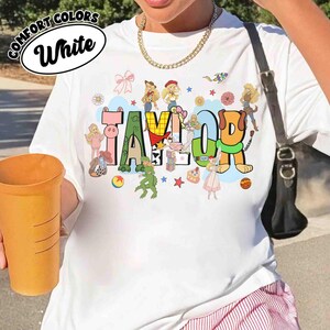 May include: A white t-shirt featuring the name TAYLOR in large, colorful, block-style letters. The letters are decorated with various cartoon characters, toys, and small icons like stars and flowers. A black shoulder bag strap is visible on the side.