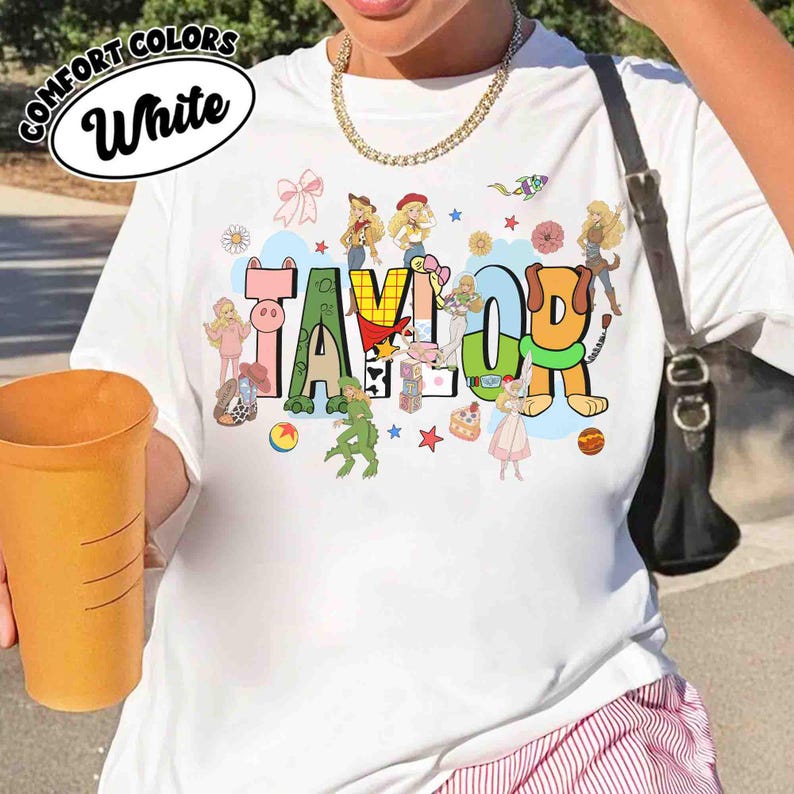 May include: A white t-shirt featuring the name TAYLOR in large, colorful, block-style letters. The letters are decorated with various cartoon characters, toys, and small icons like stars and flowers. A black shoulder bag strap is visible on the side.
