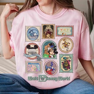 Puede incluir: Camiseta rosa claro con una colecci&oacute;n de dise&ntilde;os de sellos postales con tem&aacute;tica de Disney. Los sellos muestran personajes y logotipos de varias propiedades de Disney, incluyendo Star Wars, Toy Story y Monsters, Inc. El texto "Walt Disney World" est&aacute; impreso en la parte inferior.