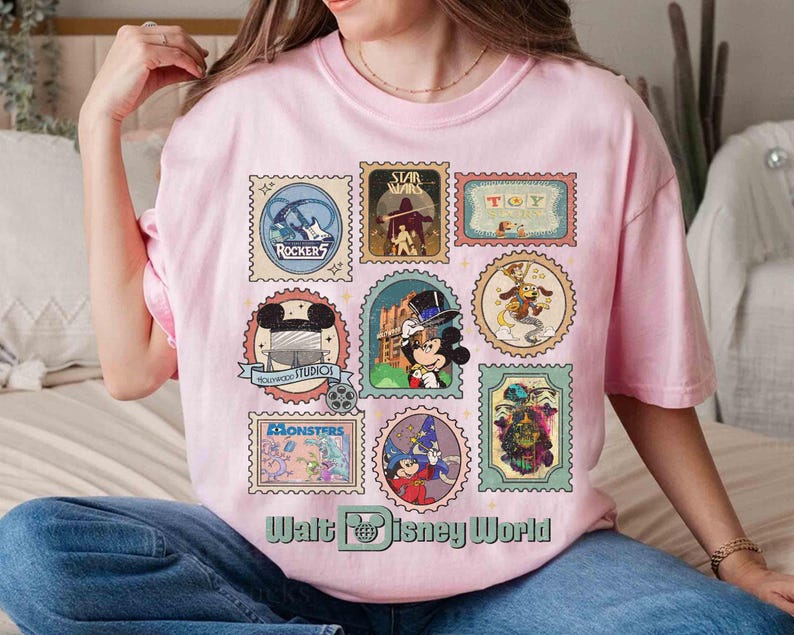 Puede incluir: Camiseta rosa claro con una colecci&oacute;n de dise&ntilde;os de sellos postales con tem&aacute;tica de Disney. Los sellos muestran personajes y logotipos de varias propiedades de Disney, incluyendo Star Wars, Toy Story y Monsters, Inc. El texto "Walt Disney World" est&aacute; impreso en la parte inferior.