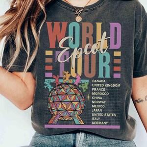 K&ouml;nnte beinhalten: Ein schwarzes T-Shirt mit einer Grafik im Vintage-Stil, die "World Epcot Tour" in bunten Buchstaben zeigt. Die Grafik zeigt auch ein stilisiertes Bild der Epcot Spaceship Earth und eine Liste der im Park vertretenen L&auml;nder.
