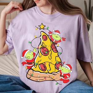 Op de afbeelding: Lichtpaarse t-shirt met een pizzapunt versierd als kerstboom. De pizza heeft pepperoni en is versierd met lichtslingers en een ster. Drie groene aliens in kerstmanpakken zijn ook afgebeeld.