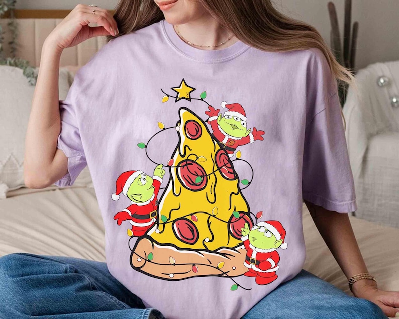 Op de afbeelding: Lichtpaarse t-shirt met een pizzapunt versierd als kerstboom. De pizza heeft pepperoni en is versierd met lichtslingers en een ster. Drie groene aliens in kerstmanpakken zijn ook afgebeeld.