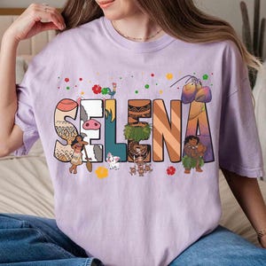 Puede incluir: Camiseta lila claro con el nombre "Selena" en letras coloridas de estilo caricatura. Cada letra est&aacute; decorada con im&aacute;genes de personajes y objetos. La camiseta es de estilo casual y c&oacute;modo.