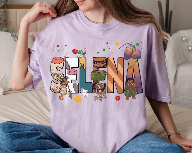 Puede incluir: Camiseta lila claro con el nombre "Selena" en letras coloridas de estilo caricatura. Cada letra est&aacute; decorada con im&aacute;genes de personajes y objetos. La camiseta es de estilo casual y c&oacute;modo.