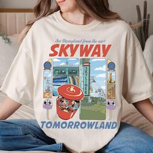 Puede incluir: Camiseta color crema con un gr&aacute;fico retro de la atracci&oacute;n Skyway de Disneyland. El dise&ntilde;o incluye las palabras "SKYWAY" y "TOMORROWLAND", junto con ilustraciones de la atracci&oacute;n y otros elementos del parque. La camiseta tiene un corte holgado.