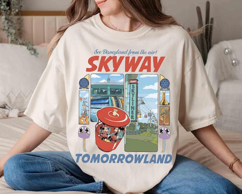 Puede incluir: Camiseta color crema con un gr&aacute;fico retro de la atracci&oacute;n Skyway de Disneyland. El dise&ntilde;o incluye las palabras "SKYWAY" y "TOMORROWLAND", junto con ilustraciones de la atracci&oacute;n y otros elementos del parque. La camiseta tiene un corte holgado.