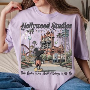 K&ouml;nnte beinhalten: Helllila T-Shirt mit einer Grafik von Hollywood Studios und Mickey Mouse. Das Design enth&auml;lt den Text "Hollywood Studios" und "That Never Was And Always Will Be". Das Bild zeigt eine Themenpark-Szene mit einer Achterbahn, Palmen und einem Wasserturm.