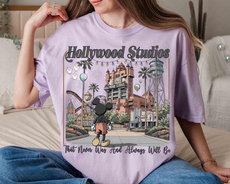 K&ouml;nnte beinhalten: Helllila T-Shirt mit einer Grafik von Hollywood Studios und Mickey Mouse. Das Design enth&auml;lt den Text "Hollywood Studios" und "That Never Was And Always Will Be". Das Bild zeigt eine Themenpark-Szene mit einer Achterbahn, Palmen und einem Wasserturm.