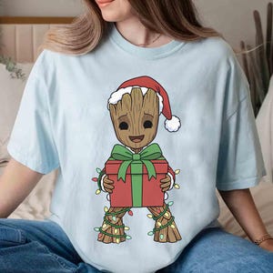 May include: Light blue t-shirt featuring a cartoon character resembling Groot from Guardians of the Galaxy, wearing a Santa hat and holding a red gift box with a green bow. The character is adorned with Christmas lights.
