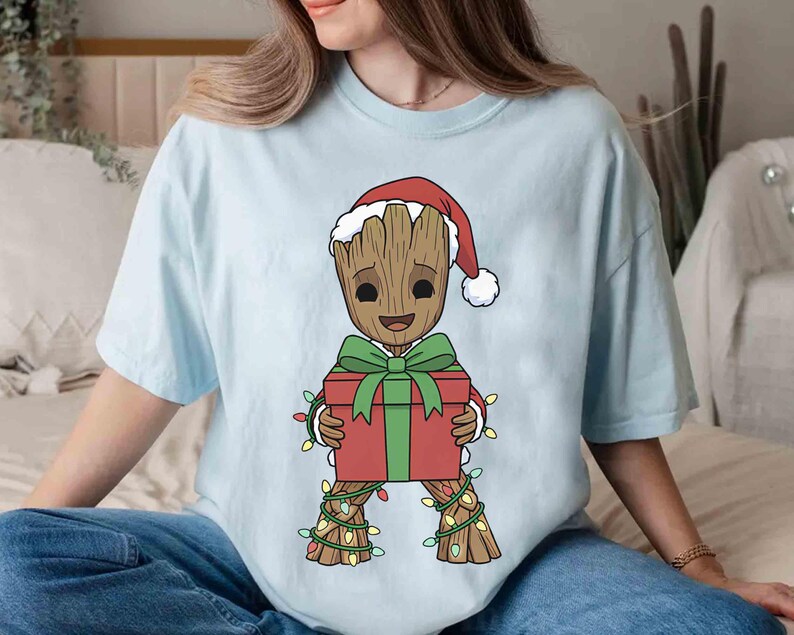 May include: Light blue t-shirt featuring a cartoon character resembling Groot from Guardians of the Galaxy, wearing a Santa hat and holding a red gift box with a green bow. The character is adorned with Christmas lights.