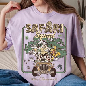 May include: A lilac t-shirt with a graphic of Disney characters in a safari jeep. The text "SAFARI Squad" is above the characters, with "Walt Disney World" below. The jeep is tan, and the characters include Mickey Mouse, Donald Duck, Goofy, and Pluto.