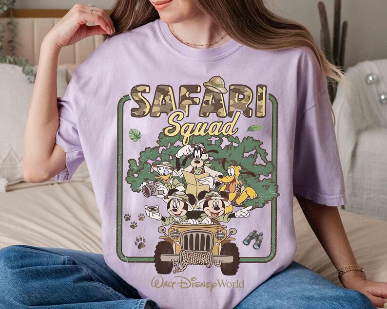 May include: A lilac t-shirt with a graphic of Disney characters in a safari jeep. The text "SAFARI Squad" is above the characters, with "Walt Disney World" below. The jeep is tan, and the characters include Mickey Mouse, Donald Duck, Goofy, and Pluto.