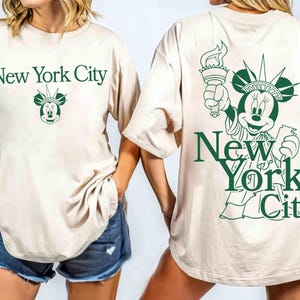 May include: Beige t-shirt with green text that says "New York City" and a Mickey Mouse head with a crown. The back of the shirt features a green illustration of Mickey Mouse as the Statue of Liberty.