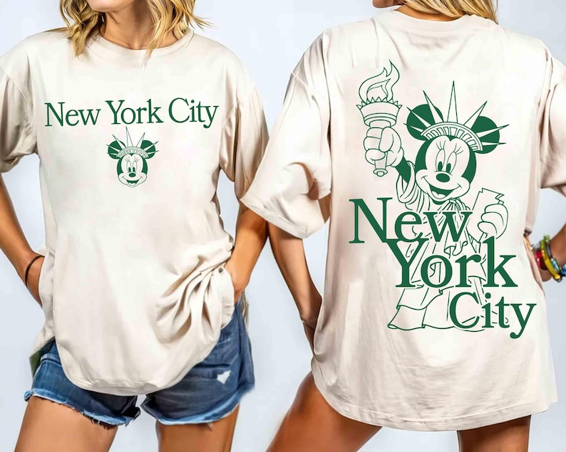 May include: Beige t-shirt with green text that says "New York City" and a Mickey Mouse head with a crown. The back of the shirt features a green illustration of Mickey Mouse as the Statue of Liberty.